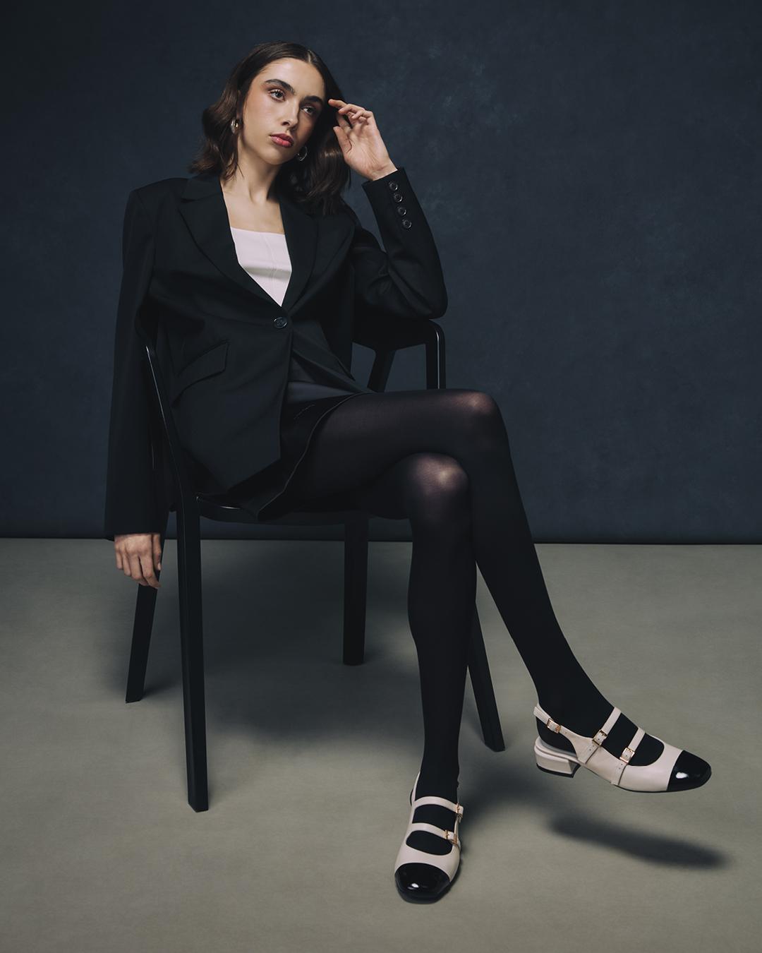 Woman sitting in front of a grey background in a womens' suit, wearing tights and black and white Mary Janes.